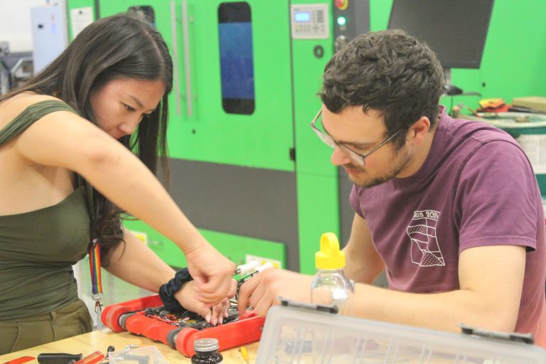 Two people work on small red robot in mechanical workshop setting.