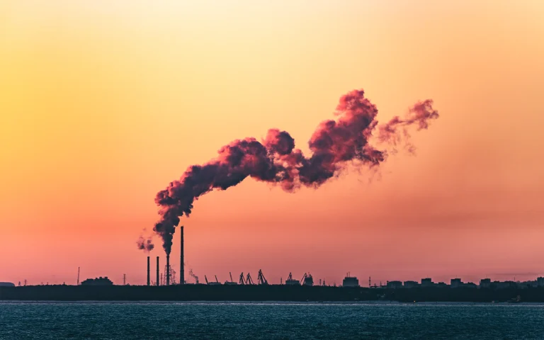 Dramatic sunset highlighting industrial smokestacks and air pollution over a city skyline by the sea.