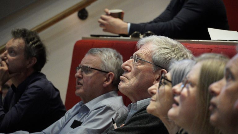 Group of men and women with grey hair sitting in a lecture theatre looking at something off camera.